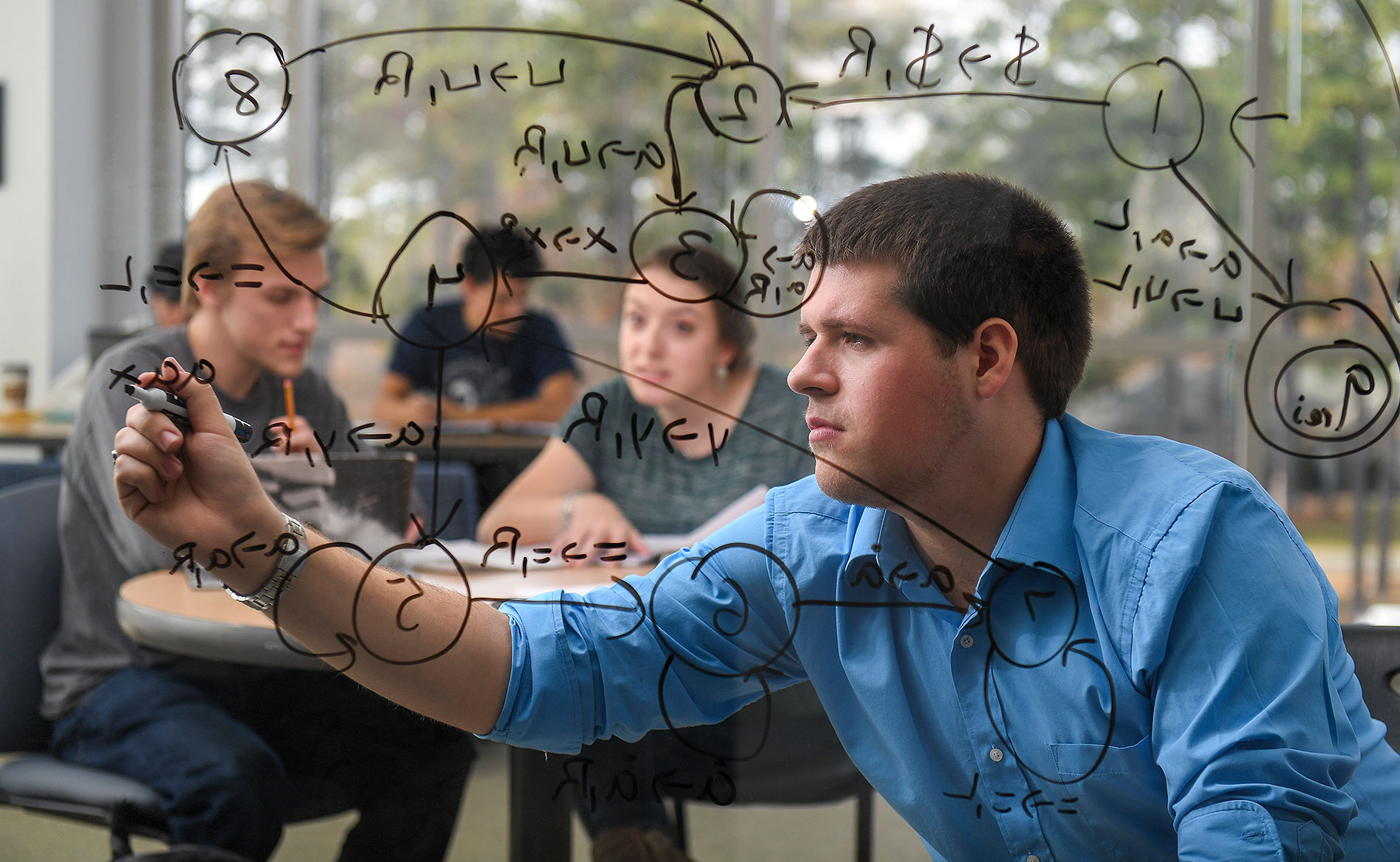 Student writing on glass with multiple students studying behind